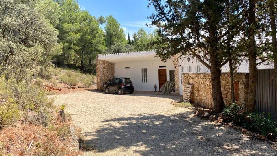 Casa de campo de una planta con fachada blanca y muros de piedra, situada al borde de un camino de grava amplio. El entorno es boscoso, con pinos y matorral mediterráneo alrededor, y se ve un porche abierto junto a una puerta de madera.