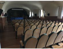 Sala de actos con techo abovedado y filas de butacas de madera orientadas hacia un pequeño escenario al fondo con mesa cubierta por un mantel azul. A los lados hay ventanas con cortinas claras, panelado de madera en las paredes y altavoces montados.