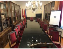 Sala de reuniones formal con una larga mesa rectangular de superficie oscura y sillas tapizadas en rojo a ambos lados, iluminada por una gran lámpara de araña. A lo largo de las paredes hay vitrinas de madera con puertas de cristal llenas de libros, banderas en un extremo y un rotafolio junto a una ventana con cortinas rojas.