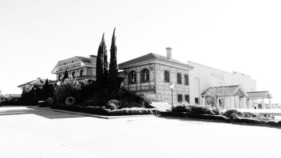 Edificio amplio de estilo tradicional con fachada de ladrillo y ventanas arqueadas, junto a un volumen anexo claro, situado al borde de una gran explanada pavimentada. Frente a la construcción hay un jardín con cipreses altos, arbustos recortados y farolas.