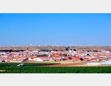Vista panorámica de un pueblo con casas bajas y tejados rojos en una llanura agrícola, con campos verdes en primer plano. En la cresta del horizonte se alinean varios molinos de viento sobre una colina bajo un cielo despejado.