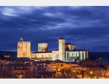 Vista nocturna de una gran catedral de piedra iluminada, con torres cuadradas almenadas y una nave con rosetón, elevándose sobre un conjunto de tejados de un pueblo. Al fondo se ven colinas oscuras y un cielo nublado.