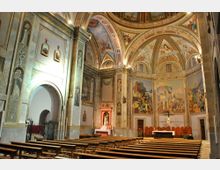 Interior de una iglesia con nave amplia y bancos de madera orientados hacia el altar, donde hay un crucifijo y un retablo lateral. Las paredes y la bóveda de arcos están decoradas con frescos y molduras ornamentales, y se ven puertas laterales y un gran arco de entrada.