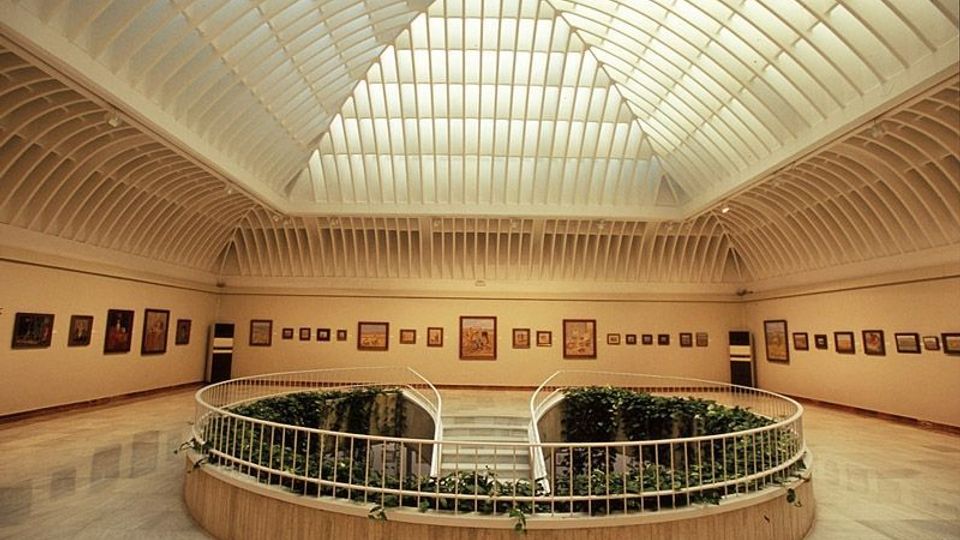 Interior de una galería o museo con un amplio techo abovedado de lucernarios y estructura blanca. En el centro hay un patio/jardín hundido con vegetación rodeado por barandillas, y en las paredes perimetrales se exhiben cuadros enmarcados.
