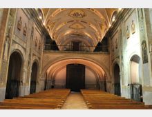 Interior de una iglesia con nave central y bancos de madera alineados hacia el fondo, flanqueada por arcos laterales y muros con frescos y relieves. El techo abovedado está decorado con pinturas, y al fondo hay una tribuna con barandilla sobre el arco principal.