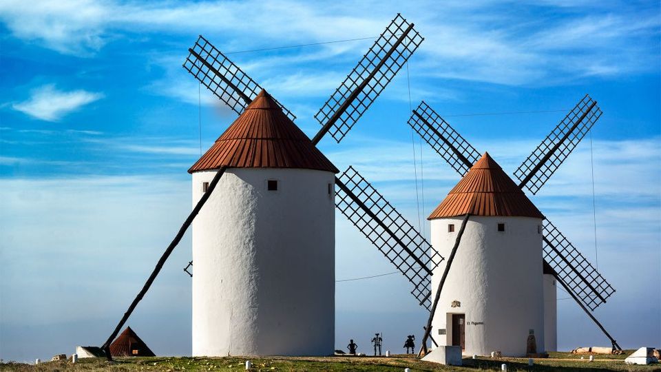 Dos molinos de viento tradicionales de paredes blancas y techos cónicos rojizos se alzan en una loma, con grandes aspas de rejilla cruzadas. El entorno es un campo abierto con vegetación baja bajo un cielo azul con nubes dispersas.