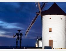 Molino de viento tradicional de cuerpo cilíndrico blanco y tejado cónico oscuro, con grandes aspas, en un entorno rural al atardecer. A la izquierda se ve una escultura metálica y al fondo aparecen otros molinos bajo un cielo azul intenso.