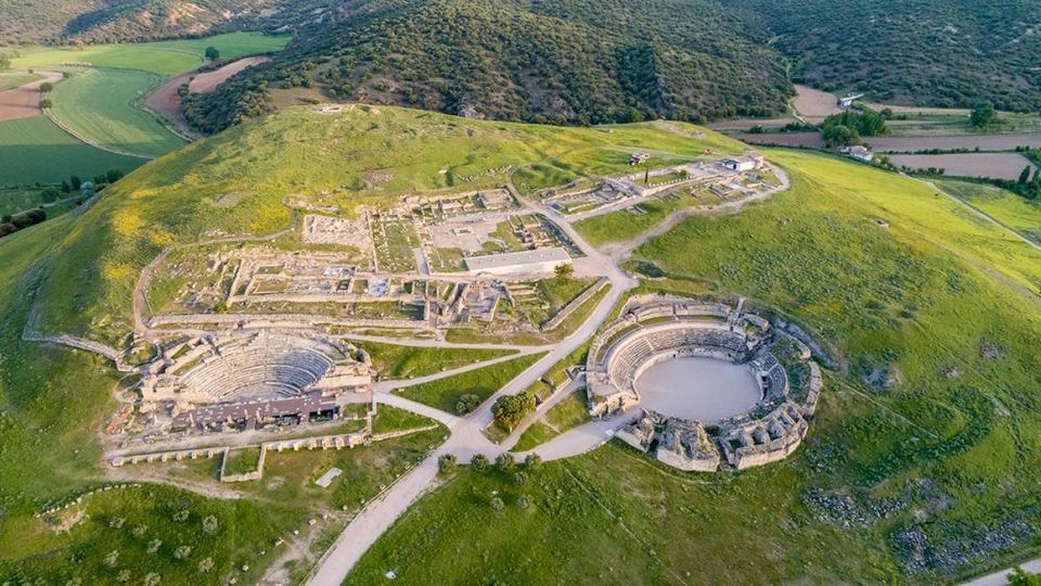 Vista aérea de un yacimiento arqueológico en una colina verde, con ruinas de edificios y caminos que conectan dos anfiteatros de piedra semicirculares. Al fondo se ven campos de cultivo y laderas boscosas alrededor del sitio.