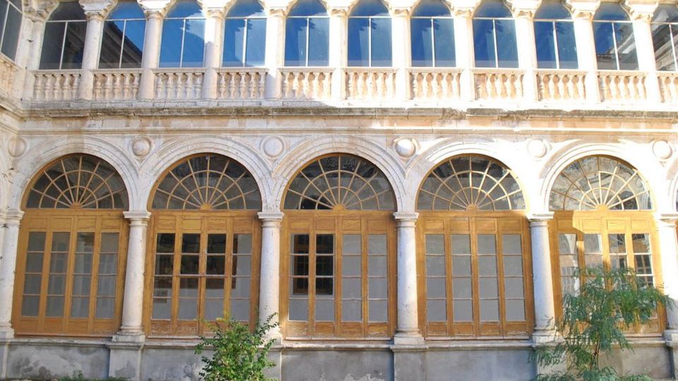 Fachada de un edificio histórico con una galería de arcos de piedra y grandes puertas-ventanas de madera con cristales, rematadas por ventanales en arco. En la parte superior se ve una logia con columnas, balaustrada y ventanas, frente a un pequeño jardín.