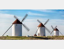 Conjunto de molinos de viento tradicionales con torres cilíndricas blancas y techos cónicos rojizos en una llanura abierta. Las aspas de celosía se recortan contra un cielo azul despejado con pocas nubes.