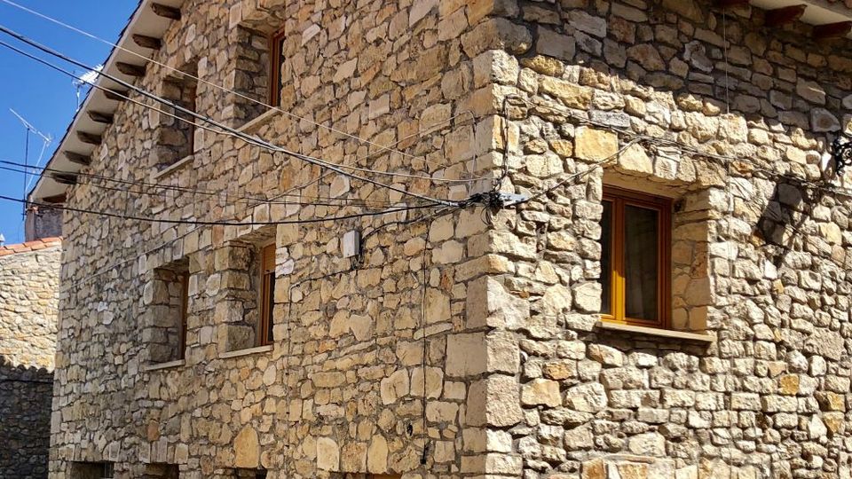 Casa tradicional de piedra en una calle estrecha, con techo de tejas, aleros con vigas de madera vistas y chimenea alta. La fachada muestra varias ventanas pequeñas con marcos de madera, una ventana con bloques de vidrio y cables aéreos cruzando frente al edificio.