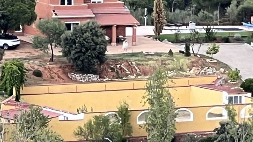 Vista de una zona residencial en ladera con una casa de ladrillo de dos plantas y paneles solares en el tejado, rodeada de árboles y vegetación. En primer plano aparece un edificio alargado de color amarillo con arcos y una carretera cercana, con campo y colinas al fondo.