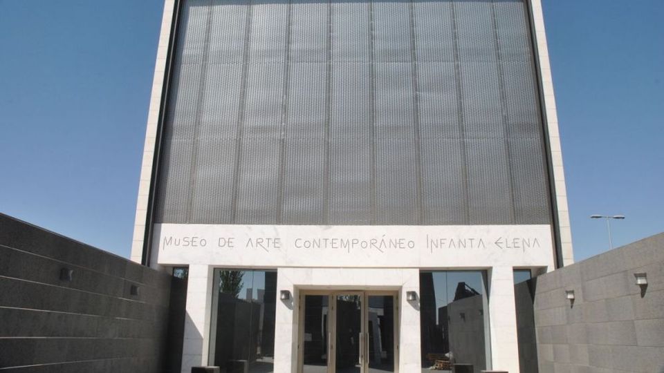 Fachada de un museo de arte contemporáneo con diseño moderno: gran panel metálico perforado en la parte superior, marco blanco rectangular y acceso central con puertas de vidrio. A ambos lados hay muros grises que forman un pasillo de entrada bajo cielo despejado.