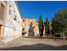 Patio amplio de tierra junto a un conjunto de edificios antiguos de ladrillo y fachadas encaladas con ventanas enrejadas y pequeños balcones. A la derecha hay árboles altos y vegetación dispersa, con bancos y bordillos de piedra bajo un cielo azul despejado.