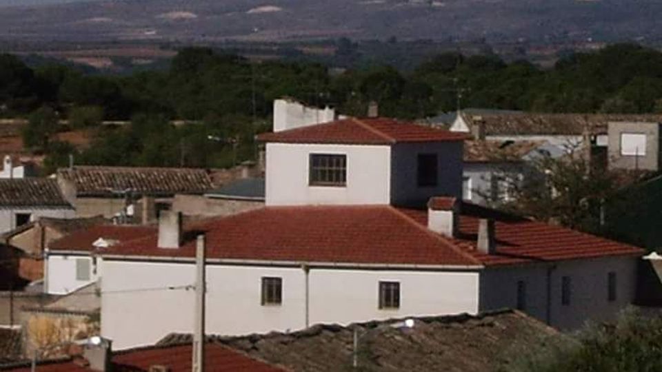 Conjunto de edificios bajos de estilo rural con fachadas blancas y tejados de teja roja, con una estructura central más alta. Alrededor se ven otras casas y árboles, con colinas y campos al fondo bajo un cielo despejado.