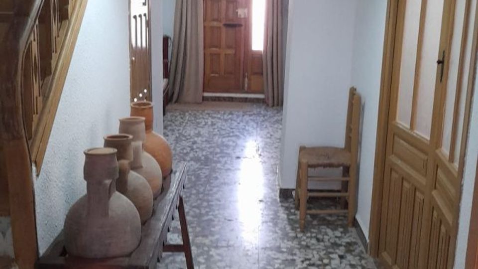 Pasillo interior de una casa con suelo de mosaico de terrazo y vigas de madera vistas en el techo, iluminado por lámparas colgantes. A la izquierda hay una escalera de madera y una mesa con jarrones de barro, y al fondo se ve una puerta de madera con cortinas.