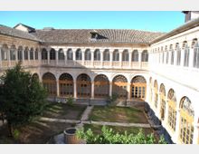Patio interior tipo claustro rodeado por una galería de dos niveles con arcos de piedra y grandes ventanales, bajo un tejado de tejas. En el centro hay parterres de césped delimitados por senderos de grava, con algunos arbustos y un pozo circular.