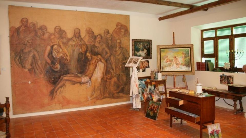 Interior de un estudio de pintura con suelo de baldosas de terracota y vigas de madera en el techo, donde destaca un gran mural en la pared. Hay caballetes con lienzos, mesas con materiales de arte y una ventana con contraventanas verdes que ilumina el espacio.