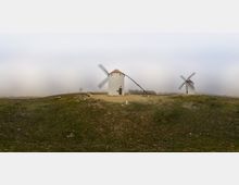Panorámica de una llanura con suaves colinas cubierta de hierba y niebla, con varios molinos de viento blancos de cuerpo cilíndrico y aspas enrejadas. En primer plano destaca un molino con techo cónico y una larga vara inclinada, sobre un camino de tierra.