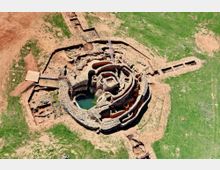 Vista aérea de un yacimiento arqueológico circular con muros concéntricos de piedra y una cavidad central con agua. Alrededor se ven zanjas de excavación y terreno mixto de tierra y pasto.