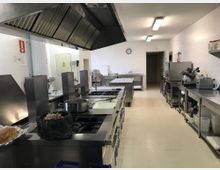 Cocina industrial con paredes alicatadas blancas y suelo claro, equipada con grandes fogones de acero inoxidable bajo una campana extractora. A ambos lados hay encimeras y estanterías metálicas con utensilios y maquinaria, iluminación fluorescente en el techo y una puerta al fondo.
