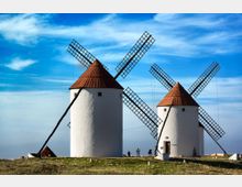 Dos molinos de viento tradicionales de paredes blancas y techos cónicos rojizos se alzan en una loma, con grandes aspas de rejilla cruzadas. El entorno es un campo abierto con vegetación baja bajo un cielo azul con nubes dispersas.