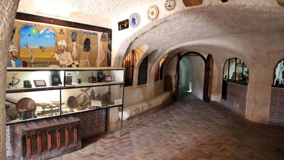 Interior de una estancia tipo museo excavada en roca, con techo abovedado y paredes blancas rugosas, suelo de ladrillo en espiga y arcos que conducen a un pasillo. A la izquierda hay vitrinas con objetos antiguos y un mural pintado, y a la derecha un mostrador con azulejos y pequeñas ventanas arqueadas con rejas.