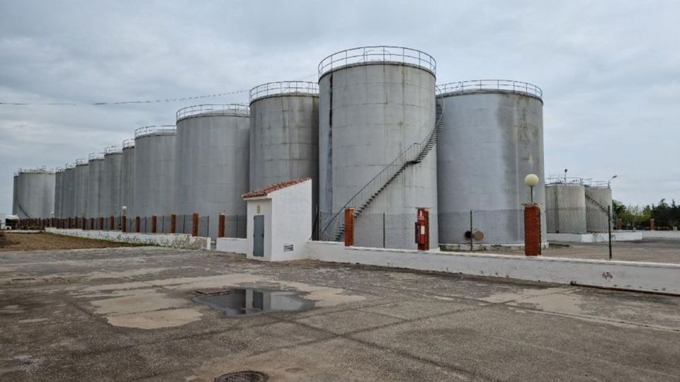 Conjunto de grandes tanques cilíndricos metálicos de almacenamiento industrial alineados, algunos con escaleras exteriores en espiral y barandillas en la parte superior. En primer plano se ve un patio de hormigón con charcos, una pequeña caseta blanca y una valla perimetral bajo un cielo nublado.