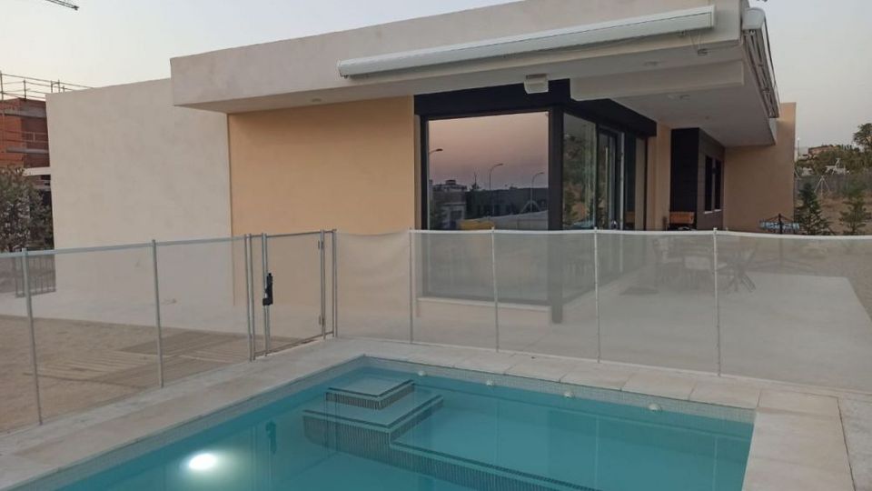 Casa moderna de una planta con techo plano y grandes ventanales, junto a una terraza con piscina rectangular. La piscina está rodeada por una valla de vidrio y pavimento claro, con vistas abiertas al entorno exterior.