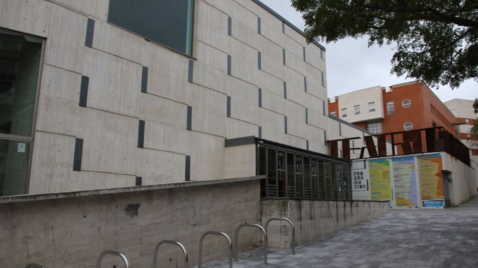Exterior de un edificio moderno de hormigón claro con ventanales rectangulares y un patrón de inserciones oscuras en la fachada. Frente a él hay una acera pavimentada con aparcabicis metálicos y, al fondo, se ven carteles en un muro y otros edificios de ladrillo junto a árboles.