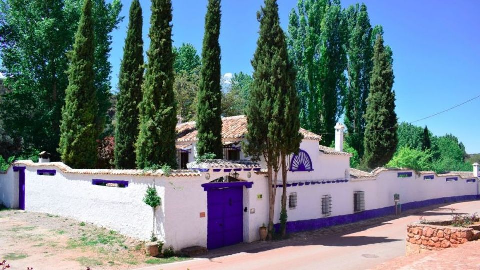 Casa rural rodeada de cipreses altos, con muros encalados y detalles en color morado, incluyendo una puerta principal y zócalos. Se ve un tejado de tejas y un camino pavimentado junto al muro bajo un cielo despejado.