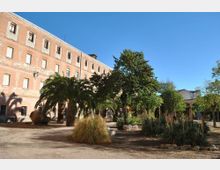 Patio ajardinado con senderos de tierra, palmeras, arbustos y árboles de sombra, junto a un gran edificio rectangular de ladrillo con varias plantas y hileras de ventanas. El entorno es abierto, con cielo despejado y vegetación distribuida en parterres.