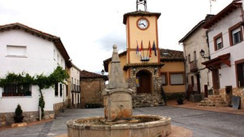 Plaza de un pueblo con una fuente circular de piedra en el centro y un monumento en forma de obelisco. Al fondo se ve un edificio con torre y reloj, y alrededor hay casas tradicionales de dos plantas con fachadas blancas y tejados de teja.