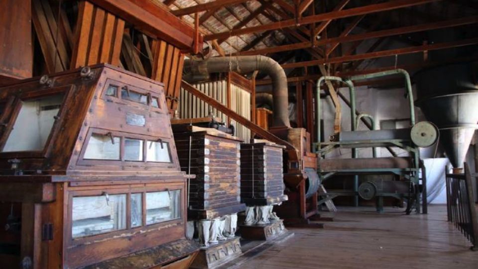 Interior de una nave industrial antigua con estructura de vigas de madera y suelo de tablas, llena de maquinaria pesada de procesamiento con paneles metálicos, conductos y grandes tolvas. Varios equipos de madera y metal se alinean a lo largo del espacio bajo un techo alto.
