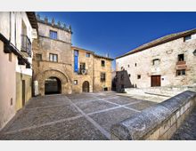 Plaza empedrada de un pueblo histórico, rodeada de edificios de piedra con arcos en la planta baja, ventanas pequeñas y tejados de teja. En primer plano se ve un murete de piedra y, al fondo, una torre con almenas bajo un cielo despejado.