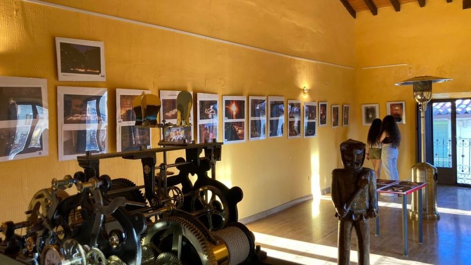 Sala de exposición con paredes amarillas y techo de vigas de madera, con una fila de fotografías enmarcadas a lo largo de la pared. En primer plano hay una gran maquinaria de engranajes metálicos y, al fondo, una escultura y mesas informativas cerca de una puerta acristalada que da al exterior.