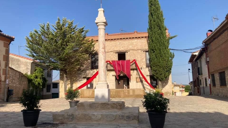 Plaza de un pueblo con pavimento de piedra y un crucero de piedra en el centro, sobre una base escalonada, coronado por una cruz de hierro. Al fondo hay una casa de piedra de dos plantas con balcones y telas rojas decorativas, flanqueada por un ciprés alto y otros árboles, con edificios bajos a los lados bajo un cielo despejado.