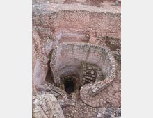Ruinas arqueológicas de piedra con un pozo circular profundo en el centro, rodeado por muros concéntricos y pasillos estrechos. Se observan escalones de piedra en espiral y barandillas metálicas rojas junto a la abertura.