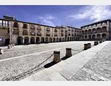 Plaza amplia empedrada con bolardos de piedra unidos por cadenas en primer plano. Al fondo, edificios históricos de piedra con soportales de arcos y balcones, y una galería porticada de dos niveles en un lateral bajo cielo despejado.