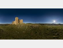 Castillo medieval de piedra con torres y murallas, situado sobre una colina cubierta de hierba en un paisaje rural abierto. Al fondo se ven colinas suaves, una carretera y postes de electricidad bajo un cielo azul con el sol bajo en el horizonte.