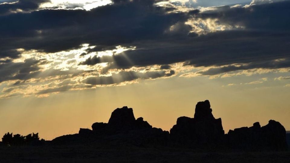 Silueta de una formación rocosa irregular en el horizonte, recortada contra un cielo de atardecer. Nubes densas cubren parcialmente el sol y dejan pasar rayos de luz hacia el paisaje oscuro.