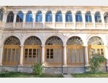 Fachada de un edificio histórico con una galería de arcos de piedra y grandes puertas-ventanas de madera con cristales, rematadas por ventanales en arco. En la parte superior se ve una logia con columnas, balaustrada y ventanas, frente a un pequeño jardín.