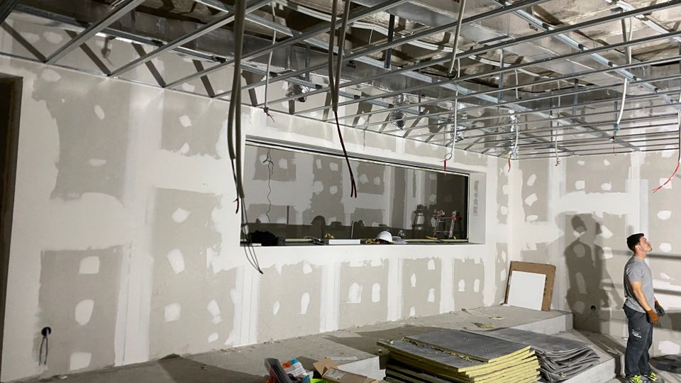 Interior de una sala en construcción con paredes de paneles de yeso con juntas selladas y estructura metálica vista para un falso techo, con cables colgando. En primer plano hay materiales y herramientas apilados, y al fondo se observa una gran abertura rectangular tipo ventana interior.
