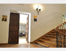 Interior de un edificio con paredes blancas, una escalera curva de madera con barandilla de hierro y una lámpara de pared. A la izquierda hay una puerta de madera abierta que da a una habitación tipo capilla, con muebles y una cruz al fondo.