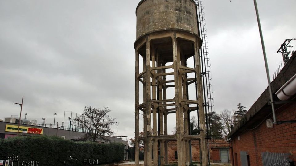 Torre elevada de un antiguo depósito de agua de hormigón, con estructura abierta de pilares y escalera vertical exterior, bajo un cielo nublado. Está en un entorno urbano-industrial junto a edificios bajos de ladrillo, setos y un área de terreno sin pavimentar.