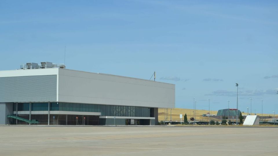 Edificio industrial moderno de gran tamaño con fachada blanca y amplias ventanas oscuras, situado junto a una extensa explanada pavimentada. Al fondo se ven farolas, una carretera y campos abiertos bajo un cielo despejado.