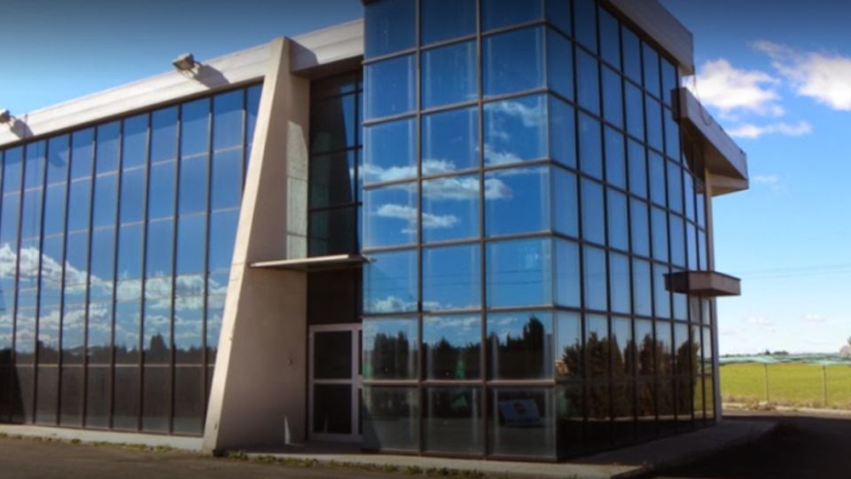 Edificio moderno de dos plantas con fachada acristalada y grandes ventanales azules, con laterales inclinados y un volumen en esquina totalmente de vidrio. Está situado en un entorno abierto junto a una carretera, con campos verdes y árboles al fondo bajo cielo despejado.