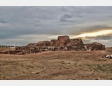 Ruinas de una fortificación de piedra y ladrillo con muros bajos y un volumen central elevado, situadas en una llanura de tierra seca. Al fondo se ve un paisaje abierto con una pasarela metálica a la izquierda y un cielo nublado con luz de atardecer.