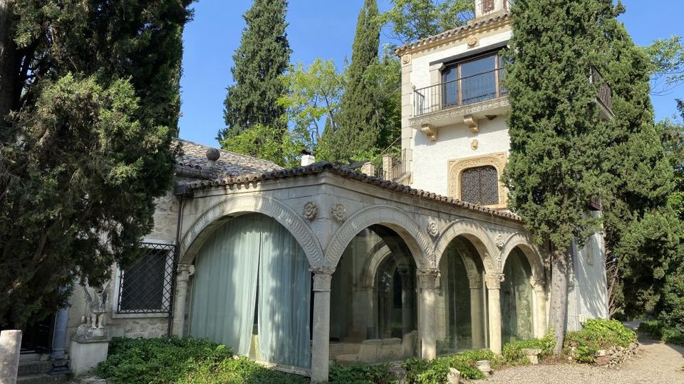 Edificio histórico de estilo mediterráneo con una galería de arcos de piedra y grandes ventanales con cortinas, junto a una torre con balcón y tejado de tejas. Está rodeado de altos cipreses y vegetación, con un camino de grava en primer plano.