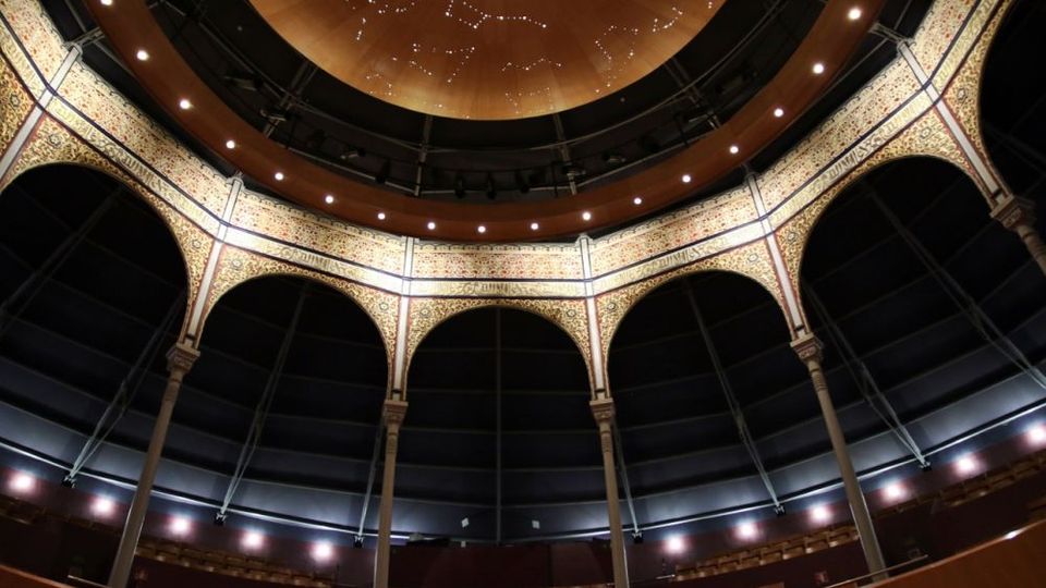 Interior de un teatro o auditorio circular con una gran cúpula central iluminada y un anillo de arcos ornamentados sostenidos por columnas. En la parte inferior se aprecian las gradas con asientos y la estructura metálica oscura de la cubierta.